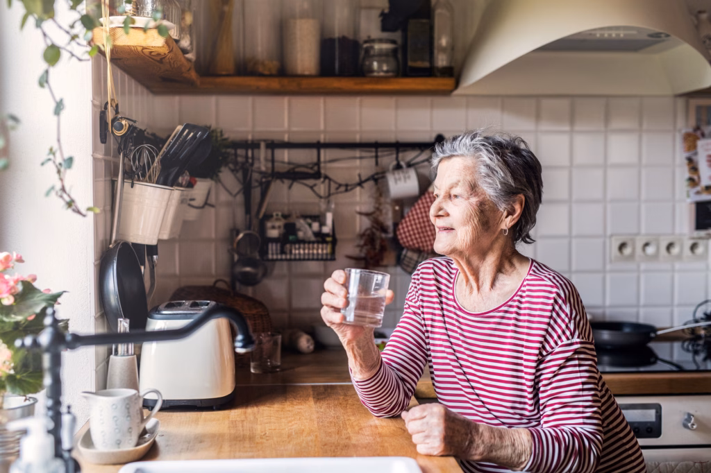 An elderly woman standing in the kitchen, holding a glass of water.
